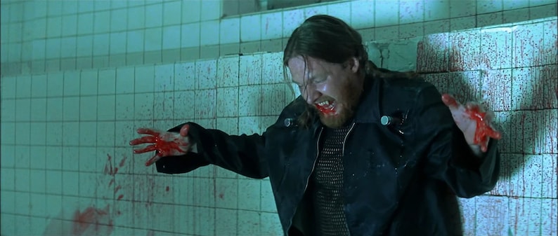 May include: A man with long hair and a beard is covered in blood. He is wearing a black leather jacket and a chainmail shirt. He is standing in a tiled room with blood splattered on the walls.