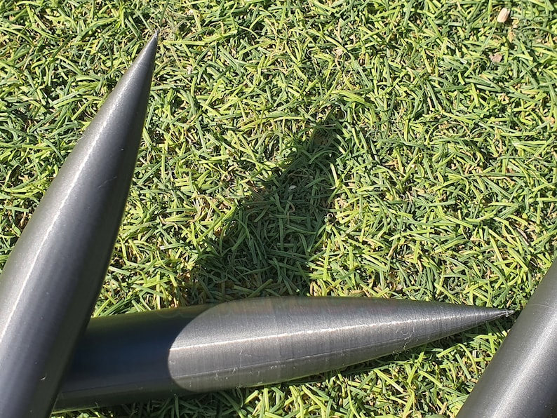 May include: Three black metal tent stakes with pointed ends, lying on a bed of green grass.