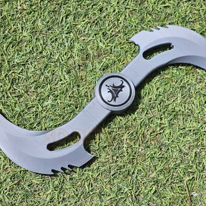 May include: A gray, double-bladed weapon with a circular emblem in the center. The blades are curved with serrated edges and a smooth, matte finish. The weapon is lying on a bed of green grass.