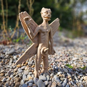 Op de afbeelding: Een beige beeldje van een gevleugelde demon met een grotesk gezicht. Het beeldje staat op een bedje van kleine, grijze stenen.