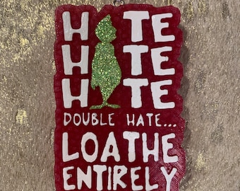 Könnte beinhalten: Roter Weihnachtsschmuck mit weißem Schriftzug: "HATE HATE HATE DOUBLE HATE... LOATHE ENTIRELY." In der Mitte befindet sich eine grüne Glitzerfigur. Der Anhänger ist rechteckig und hat eine weiße Schnur zum Aufhängen.