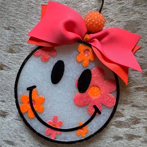 May include: A white smiley face car air freshener with a pink and orange flower design. The air freshener has a black string for hanging and a pink bow with a textured orange ball on top.