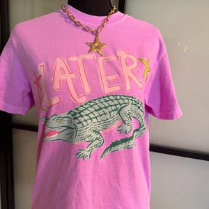 May include: A light purple t-shirt with the word "LATER" in pink and green, and a green and gray alligator graphic. A gold chain necklace with a star pendant is worn on the shirt.