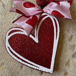 May include: A red glitter heart with a white outline, decorated with a pink and red bow with a sparkly red embellishment.