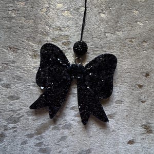 Peut inclure: Un n&oelig;ud en paillettes noires avec une perle noire au sommet, suspendu &agrave; une ficelle noire.