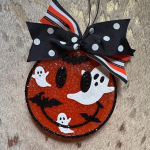 May include: A Halloween-themed air freshener with a smiling jack-o'-lantern face, white ghost and bat accents, and a black and white polka dot bow. The air freshener is round and orange with a black glitter border.