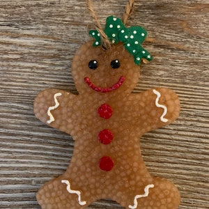 May include: A gingerbread person Christmas ornament with a green and white polka dot bow. The gingerbread person has a smiling face with red cheeks and three red buttons.