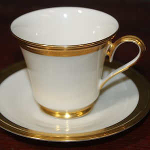 Könnte beinhalten: Eine weiße Porzellan-Teetasse und -Untertasse mit goldener Verzierung. Die Tasse hat eine abgerundete Form und einen Henkel. Die Untertasse hat einen flachen Boden und einen erhöhten Rand.