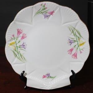 Op de afbeelding: Witte keramische serveerschaal met een gegolfde rand en een gouden rand. De schaal heeft een bloemenmotief met roze, paarse en gele bloemen.