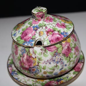 May include: A floral-patterned ceramic sugar bowl with a lid and a small matching saucer. The design features pink roses, blue and yellow flowers, and green leaves on a white background. The edges are trimmed with gold.