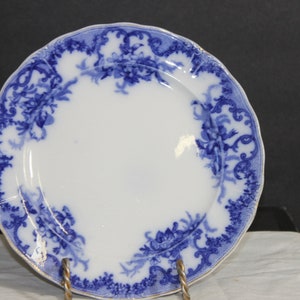 May include: A white ceramic plate with a blue floral design around the rim. The plate has a scalloped edge and a gold trim. The blue pattern features flowers and leaves. The plate is displayed on a metal stand.