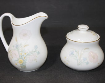 May include: A white ceramic creamer and sugar bowl set. The pitcher has a curved handle and a gold rim. The sugar bowl has a lid with a small knob. Both pieces are decorated with a floral pattern in pastel colors.