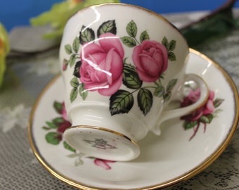 Puede incluir: Un juego de taza y platillo blanco con un patrón floral de rosas rosas. La taza tiene un borde dorado y el platillo tiene una banda dorada alrededor del borde.