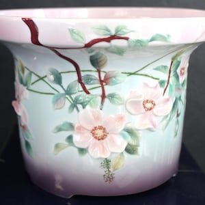 May include: A ceramic planter featuring a light blue and purple gradient base with raised, three-dimensional pink flowers, green leaves, and dark brown vine accents. The wide rim is pale pink, and the exterior surface displays a delicate floral motif.