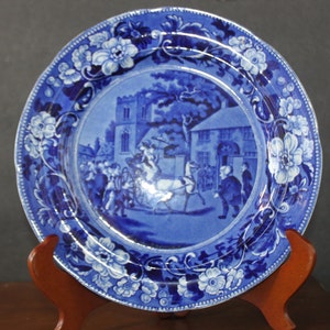 Peut inclure: Une assiette en céramique bleue et blanche avec une bordure florale et une scène de calèche devant une église et des maisons. L'assiette est sur un support en bois.