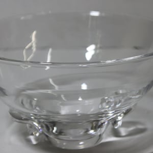 May include: A clear glass bowl with a rounded bottom and three small feet. The bowl is empty and has a smooth, reflective surface.