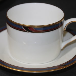 May include: A white teacup and saucer set with a gold trim and a decorative band of blue, red, and black. The teacup has a curved handle and sits on a matching saucer, creating a classic tea set.