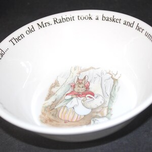 May include: A white ceramic bowl featuring a central illustration of a rabbit wearing a red shawl and bonnet, carrying a basket and umbrella. The inner rim is inscribed with the text: Then old Mrs. Rabbit took a basket and her umbrella, and