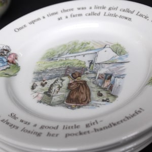 May include: A white ceramic plate featuring a central illustration of a farm scene with a child and a cat. The rim displays the text: Once upon a time there was a little girl called Lucie, who lived at a farm called Little-town. She was a good little girl—only she was always losing her pocket-handkerchiefs!