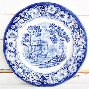 May include: A blue and white ceramic plate with a floral design around the edge. The central image depicts a landscape scene with a building, trees, and figures. The plate has a scalloped edge and is set against a white background.