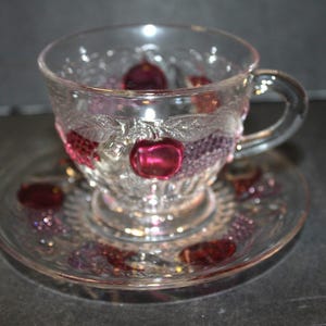 May include: A clear glass teacup and saucer set. The cup and saucer have a decorative embossed design with red and purple accents. The cup features a handle and is filled with red glass beads. The saucer also has red glass beads.