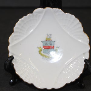 May include: White, scalloped-edge dish with a gold trim. The center features a colorful crest with a red stripe and the word "PRECIUS" in a yellow banner. The dish is displayed on a black stand.