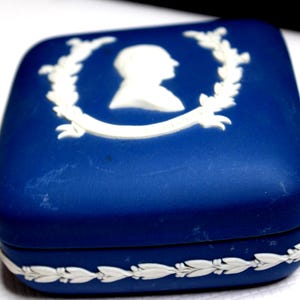May include: A square blue trinket box with rounded corners, featuring a white relief cameo of a profile portrait surrounded by a laurel wreath on the lid and a continuous white leaf garland pattern along the bottom edge of the base.
