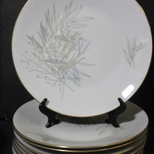 May include: A stack of white ceramic plates with a gold rim and a bamboo leaf design in gray and yellow. The plates are displayed on a black stand, showcasing the elegant design.