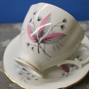 May include: A white teacup and saucer with a floral design featuring pink and grey leaves. The cup has a gold rim and the saucer has a gold band around the edge.