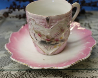 May include: A pink and white porcelain teacup and saucer set with a floral design. The cup has a heart-shaped cutout and the saucer has a scalloped edge.