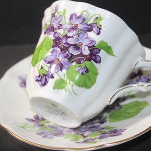 Op de afbeelding: Witte theekop en schotel set versierd met paarse viooltjes en groene bladeren. De kop heeft een gebogen handvat en een gouden rand. De schotel heeft ook een gouden rand. Een vintage theeservies.