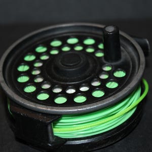May include: A black fly fishing reel with a bright green fishing line. The reel has a circular design with multiple holes and a black handle. The fishing line is neatly wound around the reel.