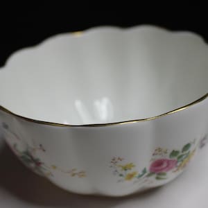 Puede incluir: Un cuenco de cerámica blanca con borde festoneado y acabado dorado. El exterior presenta un delicado patrón floral con pequeñas rosas rosadas, flores amarillas y hojas verdes dispersas por la superficie lisa y curva del cuenco.