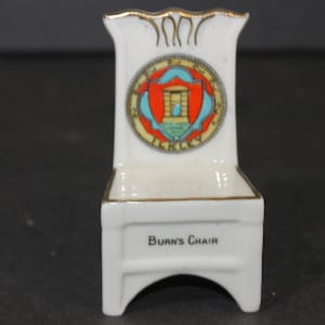 May include: A white porcelain miniature chair with gold accents. The chair has a crest with a red shield and blue and gold details. The chair is labeled "Burn's Chair".