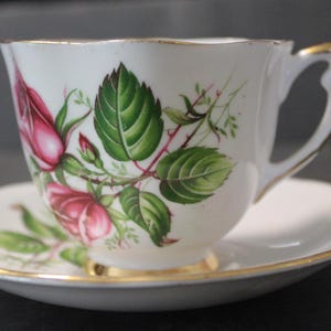 May include: White teacup and saucer set with a floral rose design. The cup and saucer have gold trim. The roses are pink with green leaves and stems. The teacup has a curved handle.