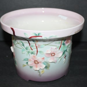 Könnte beinhalten: Ein weißer Keramiktopf mit rosa Rand und floralem Design. Der Topf zeigt rosa Blüten mit grünen Blättern und roten Stielen. Der Topf hat einen Farbverlauf, der von hellblau nach rosa verläuft.