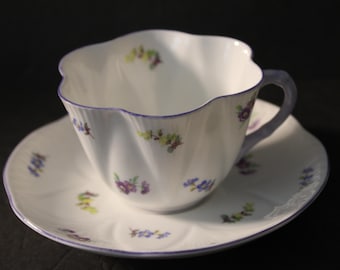 Peut inclure: Un service à thé blanc composé d'une tasse et d'une soucoupe, avec un bord festonné et un motif floral délicat en violet, jaune et bleu. La tasse a une anse assortie et une bordure violette.