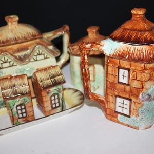 Op de afbeelding: Een keramische theeset met een cottage-ontwerp. De set bevat een theepot, een melkkan, een suikerpot en een dienblad met zout- en peperstrooiers. De stukken zijn geschilderd in bruin-, beige- en groentinten.