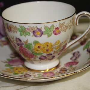 Könnte beinhalten: Eine weiße Teetasse und Untertasse mit einem floralen Muster in den Farben Rosa, Lila, Gelb und Grün. Tasse und Untertasse haben einen goldenen Rand.