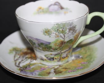 May include: A vintage teacup and saucer set with a green handle and a delicate floral design featuring a landscape scene with trees and a mountain in the background.