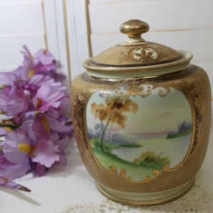 May include: A vintage ceramic cookie jar with a gold lid and a hand-painted scene of a lake and trees. The jar has a gold and white floral pattern.