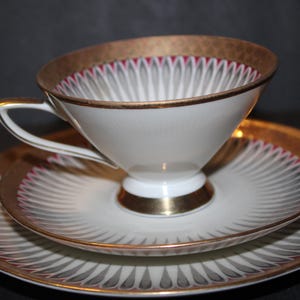 May include: A white teacup, saucer, and plate set with gold trim and a decorative pattern. The cup and plates have a gold rim and a design of gray and red lines radiating from the center. The set is on a dark background.