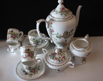 Könnte beinhalten: Weißes Porzellan-Teeservice mit Blumen- und Landschaftsdesign. Das Set beinhaltet eine Teekanne, eine Zuckerdose, einen Sahnekännchen, Tassen und Untertassen. Das Design zeigt rosa, rote und grüne Blumen mit goldenem Rand. Ein klassisches Teeservice.