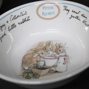 Puede incluir: Un cuenco de cerámica blanca con una ilustración de Peter Rabbit en el centro que muestra tres conejos, una cesta y una jarra. El borde incluye el texto Peter Rabbit y las frases Flopsy, Mopsy and Cottontail were good little rabbits y They went down the lane to gather blackberries.
