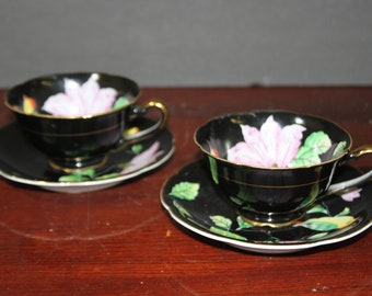 Puede incluir: Dos tazas de té negras y platillos con un diseño floral. Las tazas y los platillos tienen un fondo negro con una flor rosa y hojas verdes.