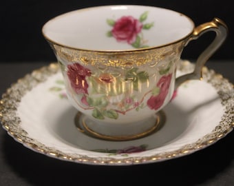 Puede incluir: Juego de taza y platillo blancos con un diseño floral de rosas rojas y verdes. La taza y el platillo tienen ribetes dorados en los bordes y el asa. Un juego de té vintage clásico.