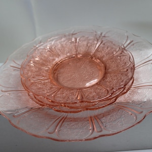 May include: A stack of three translucent pink glass plates featuring a delicate embossed floral pattern and scalloped edges. The plates are circular with a smooth center and a textured rim, showcasing a light rosy hue throughout the glass material.