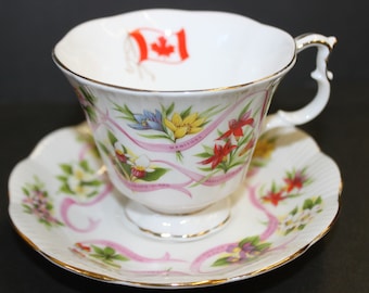 May include: White teacup and saucer set with a gold trim. The teacup features a Canadian flag and floral designs with the word "Manitoba" on a pink ribbon. The saucer has matching floral and ribbon patterns.
