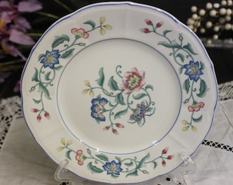 May include: White ceramic plate with a scalloped edge and a blue rim. The plate features a floral design with blue, pink, yellow, and green accents. The plate is displayed on a clear stand.