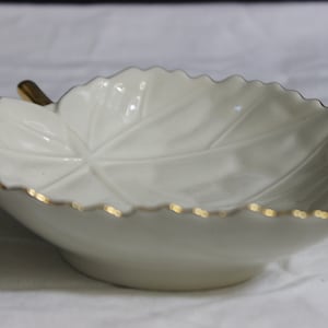 May include: A white, leaf-shaped ceramic dish with a gold-colored rim and stem. The dish has a detailed leaf vein pattern and is likely used for serving small items or as a decorative piece.